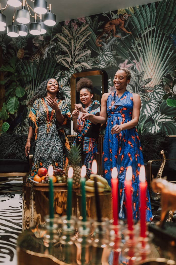 Three women celebrating Kwanzaa with traditional African attire and kinara candles indoors.